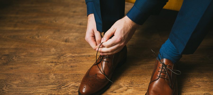 A man in a blue suit ties up shoelaces on brown leather shoes brogues on a wooden parquet background